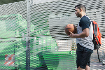 Attractive basketball player walks beside a metal wall carrying his backpack and his orange ball
