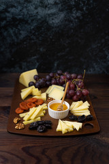 A large board with sliced different types of cheeses with honey, grapes, nuts and dried fruits. A set of cheese plates for a party and guests on a dark background with copy space.