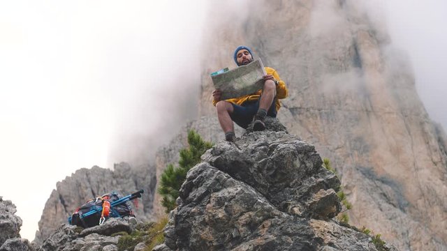 Young man hiker sitting on stone mountain reading map, with cloudy sky and fog. Yellow jacket, backpack, black beard and beanie. Traveling dolomites, Italy.