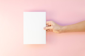Women hand hold white template paper or magazine on pink background. Mockup flat lay with copy space