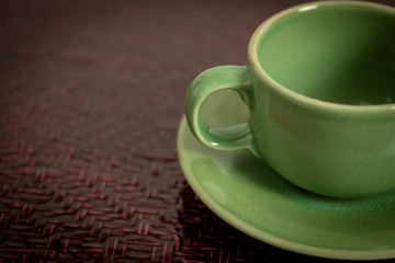 Empty glazed green coffee cup with saucer on red dark woven textured background.