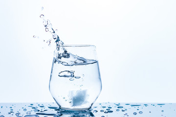 motion of ice cubes inside a drinking glass of water with big splashes and bubbles, water drops on a mirror dark blue surface, transparent blue backlight, fresh cooled drink  