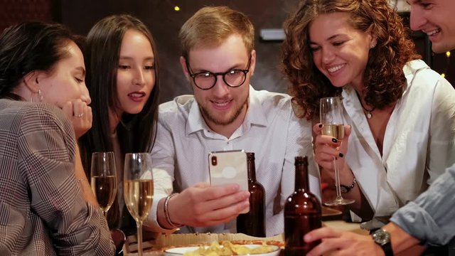 Portrait shot of group of happy hipster students watching video, photo on the smart phone together while sitting at the wooden table in the modern bar, pub.