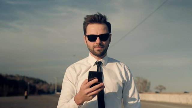 Walking And Using Mobile Phone On.Handsome Businessman Chatting On Mobile Phone With Friend  Sending Message On Social Network.Holding Using Smartphone Outdoors.Freelancer Man Mobile Working Outdoors.
