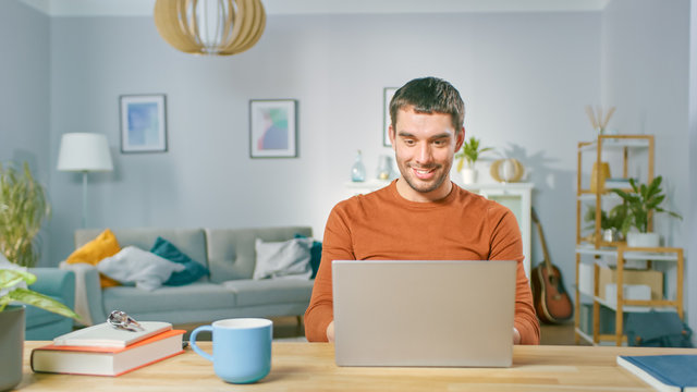 Portrait of Handsome Smiling Man Working on Laptop, Sitting at His Wooden Desk at Home. Man Browsing Through Internet, Working on Notebook from His Living Room.
