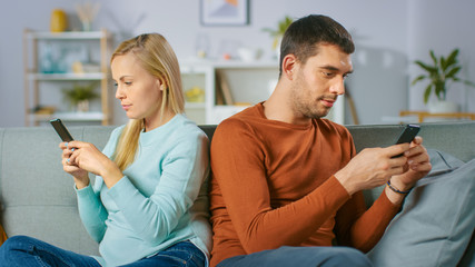 Alienation / Quarrel Concept: At Home on a Sofa Young Couple Sitting Apart, Facing Different Directions and Using Smartphones. 
