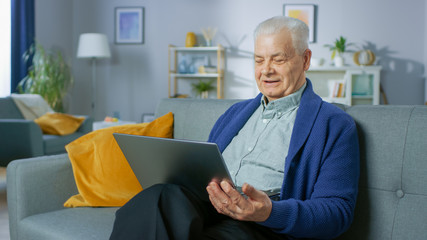 Progressive Senior Man at Home Sitting on Sofa Uses Laptop Computer. Full of Life Elderly Man Relaxing at Home, Reading News on the Internet.