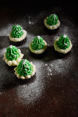 Obraz na płótnie Canvas green cakes with the addition of white cream in the form of Christmas trees on a dark textured background are in different order sprinkled with powdered sugar