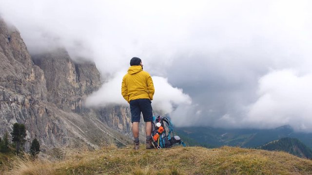 Young man hiker standing on mountain viewpoint with cloudy sky and fog. Yellow jacket, backpack, black beard and beanie. Traveling Dolomites, Italy.