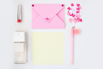 Flat lay: yellow paper, pink envelope with rhinestones, pen, pompom, perfume, lipstick, confetti hearts. Making postcard in envelope for Valentine's Day. Do it yourself. Photo from the series