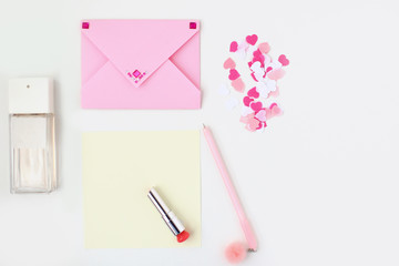 Flat lay: yellow paper, pink envelope with rhinestones, pen, pompom, perfume, lipstick, confetti hearts. Making postcard in envelope for Valentine's Day. Do it yourself. Photo from the series