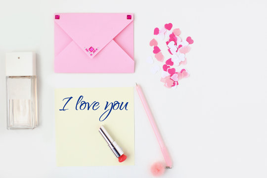 Flat lay: yellow paper, pink envelope, pen, pompom, perfume, lipstick, confetti hearts, lip print, i love you text. Making postcard in envelope for Valentine's Day.Do it yourself.Photo from the series