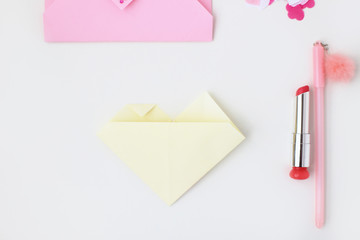 Flat lay: pink envelope, lipstick, pen, confetti hearts, yellow paper folded in the shape of heart in the origami technique. Making postcard for Valentine's Day. Do it yourself. Photo from the series