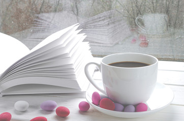 Cozy concept: a white cup of coffee stands on a white wooden table next to a white open book and colorful candy against the background of a window with raindrops