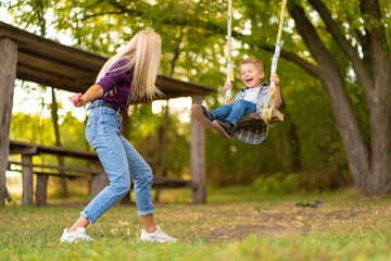 Young blonde mom shakes her little son on a swing in a green park. Happy childhood.