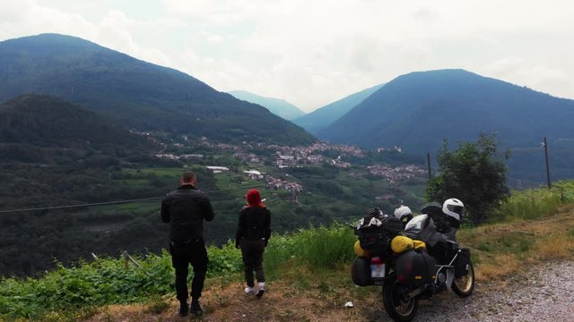 Couple of the motorcycle travelers standing with adventure motorbike and look to distance, mountain city on background, back view, Europe Faver Italy Dolomites, travel tourism concept.
