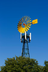 Yellow windmill in a telephone tower behind a tree on blue sky background. Alternative and renewable energy. Care of the environment and the planet.