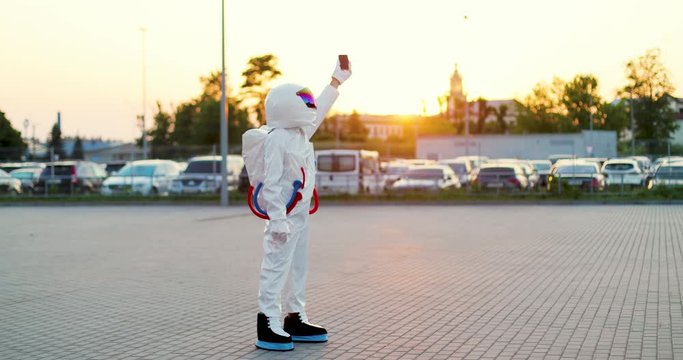Funny man astronaut in the white cosmic costume and armor walking at the street near the parking with a smartphone in hand and trying to catch mobile phone connection from the space. Outdoors.