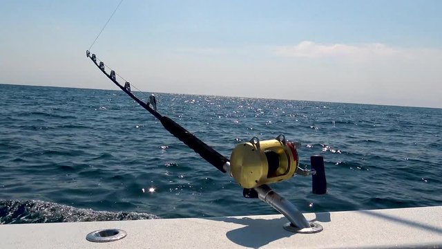 Fishing rod and reel on the side of a boat as the blue ocean passes by on a bright, sunny day.