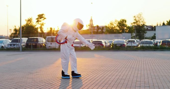 Funny astronaut in the costume and armor walking a moonwalk and moving like dancing at the cars parking on the sunset outside.