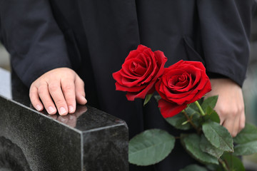 Woman with red roses near black granite tombstone outdoors, closeup. Funeral ceremony
