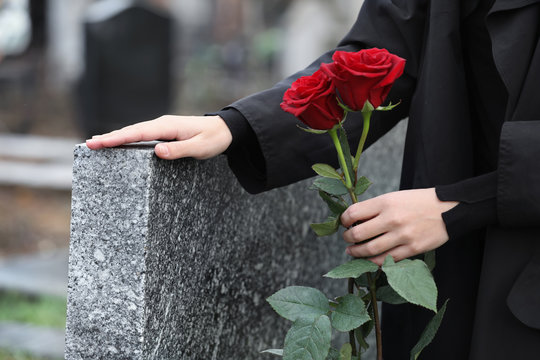 Woman with red roses near grey granite tombstone outdoors, closeup. Funeral ceremony