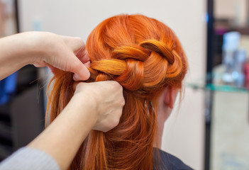Fototapeta premium Beautiful girl with long redhair, hairdresser weaves a braid, close-up in a beauty salon. Professional hair care and creating hairstyles.