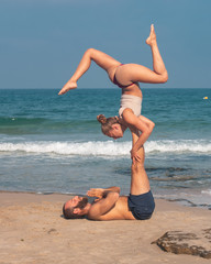 Muscular man and fit young woman training together in Acro Yoga. Morning sun lighting the couple and the mediterranean beach background.