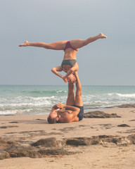 Muscular man and fit young woman training together in Acro Yoga. Morning sun lighting the couple and the mediterranean beach background.