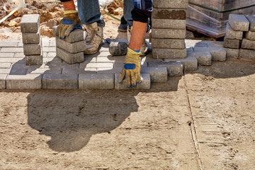 A worker lays paving slabs on a prepared flat sandy platform on the sidewalk on a bright sunny day.