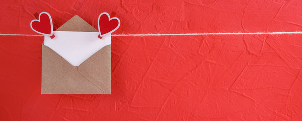 Banner with empty sheet of paper in envelope hangs on a rope attached with heart-shaped clothespins on a red background. concept of Valentine's day, anniversary, mother's day or birthday greeting