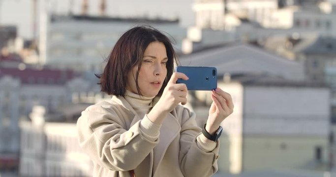 Close-up of brunette Caucasian woman with brown eyes standing on city street and taking photos. Businesslady using smartphone to photograph skyscrapers. Lifestyle, hobby. Cinema 4k ProRes HQ.
