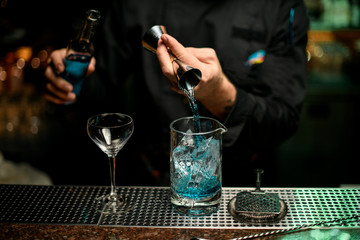 Male bartender pouring a blue alcoholic drink from the jigger to a measuring cup holding a glass bottle