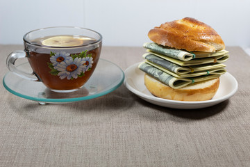 On a gray background is a light breakfast - a cup of tea with lemon and a sandwich with a bundle of money.