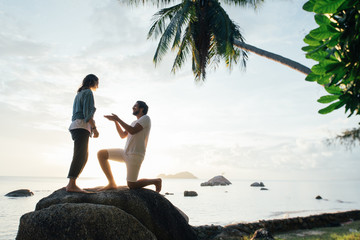 The guy makes an offer to the girl on a large stone on the seashore at sunset.