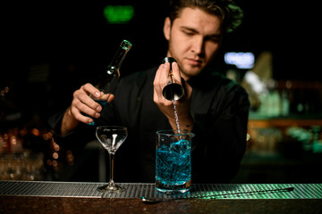 Professional male bartender pouring a blue alcoholic liquor from the jigger to a measuring cup