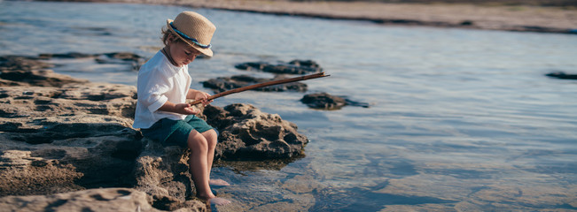 A little boy in a stylish hat sits on a stone and plays with a wand and water.