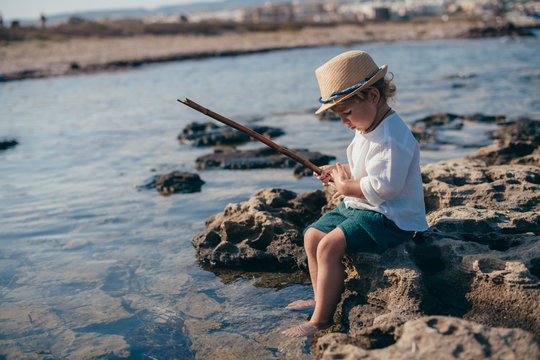 A little boy in a stylish hat sits on a stone and plays with a wand and water.