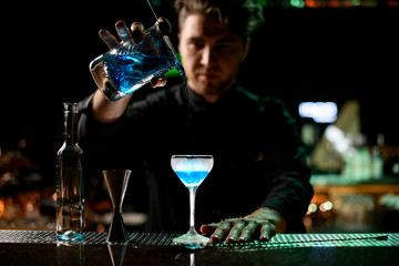 Professional male bartender pouring a blue alcoholic drink from the measuring cup to the glass through the strainer filter
