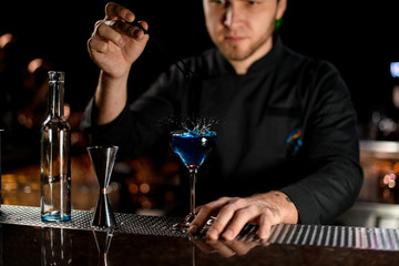 Professional male bartender throwing to the blue alcoholic drink one berry with tweezers
