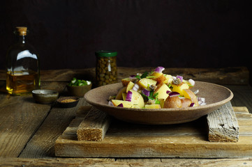 Boiled potato salad with Dijon mustard, spices, red onions and herbs. Capers, olive oil, parsley in the background, on a wooden background. Horizontal arrangement, close-up.