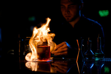 Professional bartender serving the red alcoholic cocktail with ice fire the lemon slice on tweezers