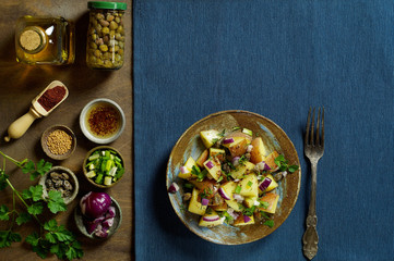 Potato salad with Dijon dressing with ingredients: capers, red and green onions, sumac seasoning, parsley, olive oil and fork. Textured blue napkin. Top view, horizontal layout, copy space.