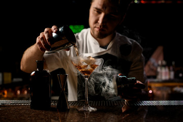 Professional bartender pouring a brown alcoholic drink from the steel shaker to the glass with the steam