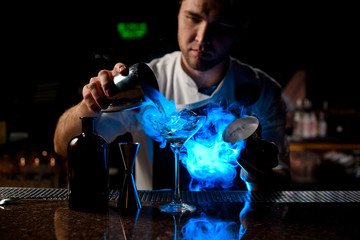 Professional male bartender pouring a brown alcoholic drink from the steel shaker to the glass under blue light