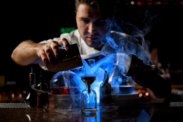 Male bartender pouring a transparent alcoholic drink from the steel shaker to the little bottle under blue light
