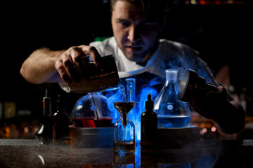 Bartender pouring a transparent alcoholic drink from the steel shaker to the little bottle under blue light