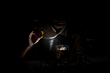 Professional male bartender spraying on the brown alcoholic drink with a orange zest juice in the dark