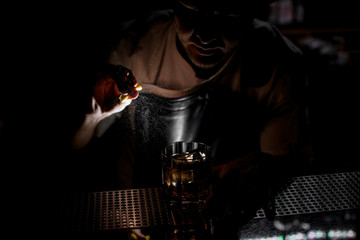 Fototapeta na wymiar Bartender spraying on the brown alcoholic drink with a orange zest juice in the dark