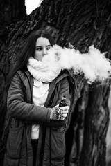 Smoking teenager in real life. Young beautiful white teeage girl in scarf smokes an cigarette near the tree on the street in the autumn. Deadly bad habit. Black and white.